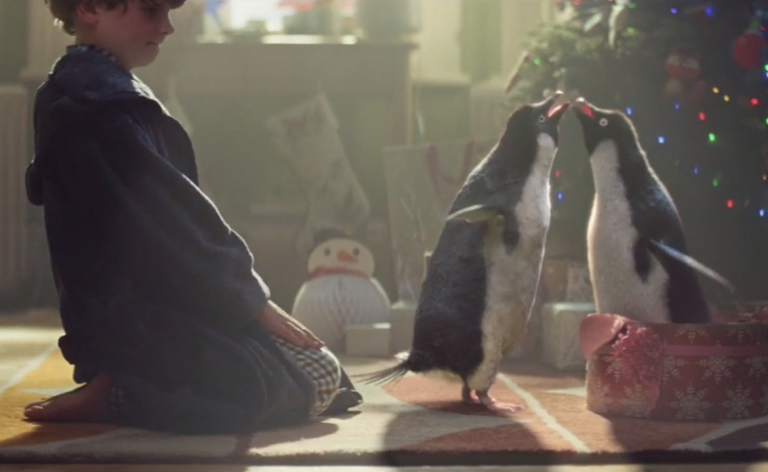 Two penguins nuzzling under a Christmas tree while a boy watches.